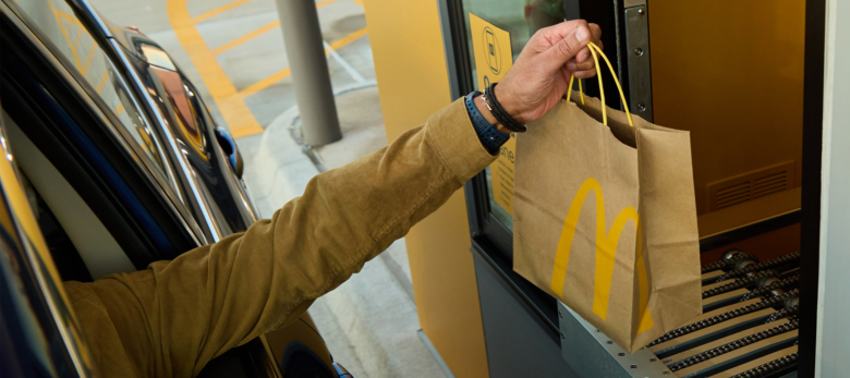 A drive-through McDonald's window showing a customer's arm through the car window grabbing a bag off an automated conveyor.